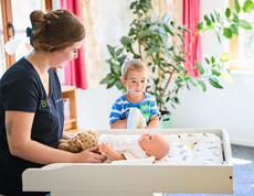 Eine Schülerin der Diplom-Sozialbetreuung Familienarbeit versorgt eine Pflegepuppe auf einem Wickeltisch. Ein Kleinkind steht dahinter und sieht gespannt zu. 