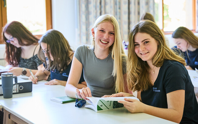 Schüler*innen sitzen im Klassenzimmer. 2 Schülerinnen lesen ein Buch über Familienarbeit und lächeln.