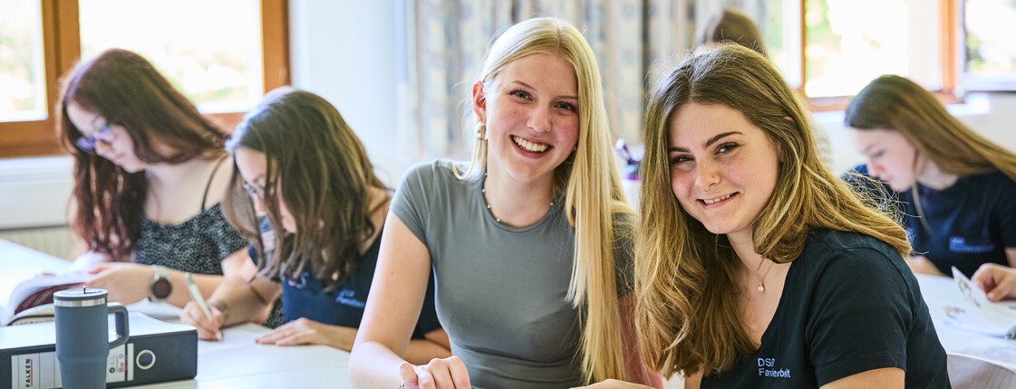 Schüler*innen sitzen im Klassenzimmer. 2 Schülerinnen lesen ein Buch über Familienarbeit und lächeln.